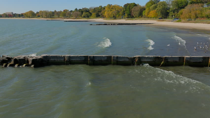 Fast-moving aerial pulling back from a pier near the beach over Lake Michigan in Sheboygan, Wisconsin. Vibrant autumn trees line the shore as gentle waves shimmer under the crisp seasonal sunlight