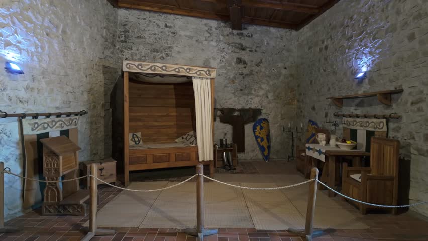 A reconstructed medieval bedroom inside Hollókő Castle, Hungary. The room features a wooden canopy bed, shields, medieval furniture, and stone walls.