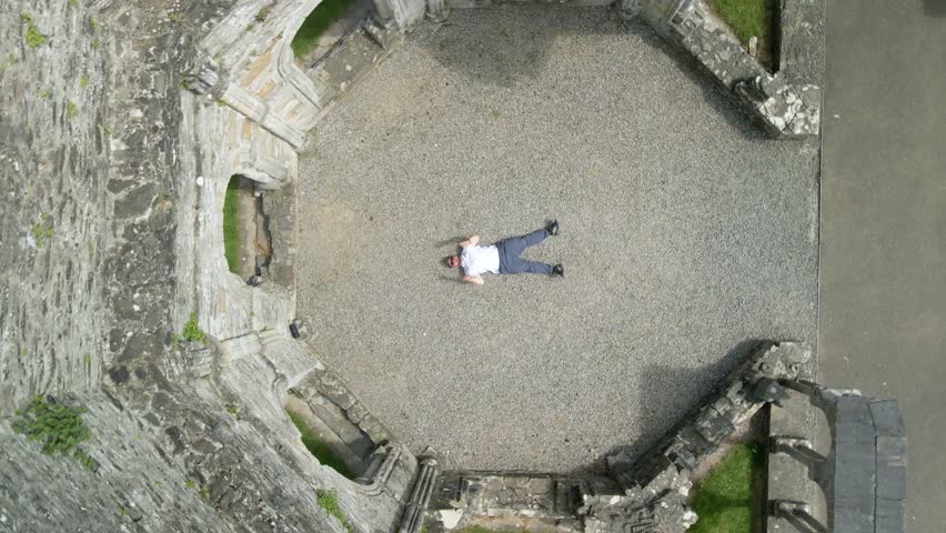 Male Rapper Lying In Ruins Of Old Mellifont Abbey In County Louth, Ireland. aerial topdown, rotating shot