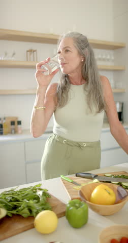 Vertical video: In kitchen, woman drinking water while preparing vegetables on countertop. Cooking, hydration, healthy, fresh, slow motion