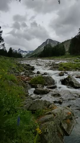 A serene mountain stream flows through a landscape of green meadows and rocky outcrops under a cloudy sky, creating a peaceful atmosphere.