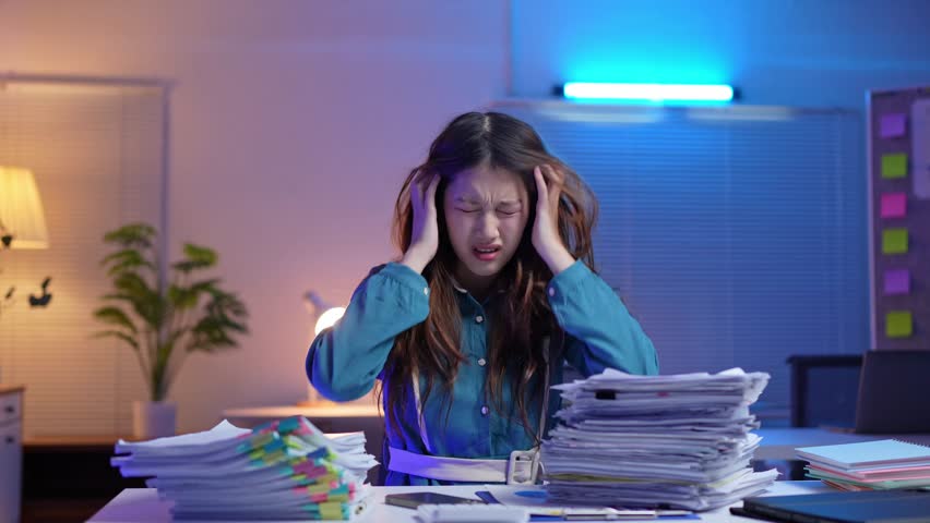 Asian office worker is extremely stressed by her workload, pulling her hair, crumpling papers, and eventually resting her head on the desk, defeated