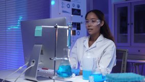 Young female scientist working on computer in modern laboratory with blue chemical liquid in beaker and test tubes, wearing lab coat and protective glasses, analyzing data and conducting research - Powered by Shutterstock - Get 15% off with code: PIKWIZARD15