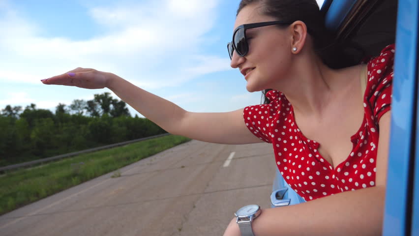 Young girl in sunglasses leaning out of vintage car window and enjoying trip. Woman looks out from moving retro auto at summer day. Travel concept. Slow motion Close up