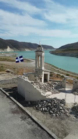 Aerial drone view of abandoned church ruin set against Cyprus’s vast Kouris Water Dam landscape, showcasing rugged terrain, serene waters, and historical architecture amidst nature.