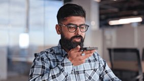 Bearded businessman using voice assistant on smartphone. Young confident male wearing glasses in plaid shirt using mobile phone indoors. Modern professional using smartphone in office setting. - Powered by Shutterstock - Get 15% off with code: PIKWIZARD15