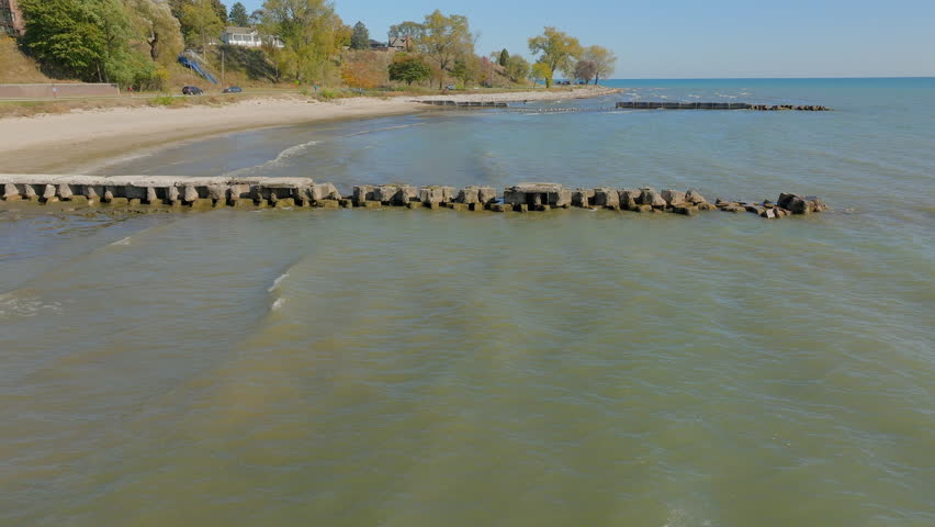 Aerial shot moving dynamically over Lake Michigan, gliding along the beach and passing over old jetties in Sheboygan, Wisconsin. Vibrant autumn trees, gentle waves, and piers.