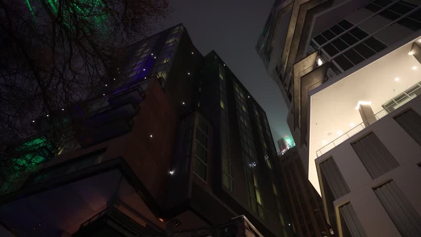 A smooth, moody night shot glides past towering buildings, their dark silhouettes punctuated by dimly glowing windows and streetlights.