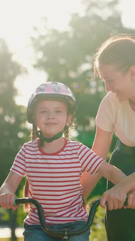 Elder sister teaches kid girl to ride bike park. Child cyclist with mom, nature. Sport family, little girl in helmet together with her mother learns to ride bicycle outdoors in summer. Family weekend