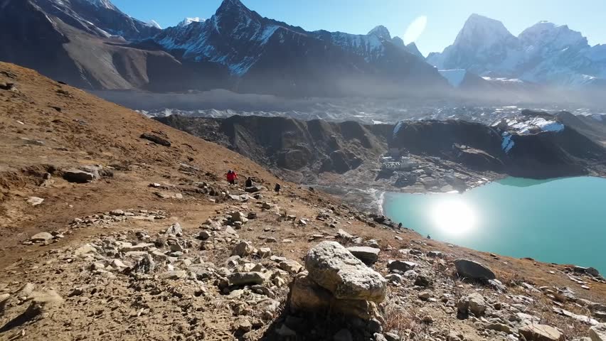 An elevated viewpoint captures the stunning beauty of Gokyo and its turquoise lake nestled amidst the Himalayas on a sunny morning, with hikers slowly ascending the surrounding slopes