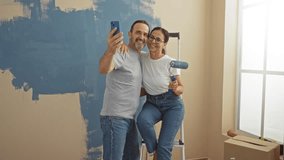 Hispanic couple smiling as they take a selfie during home renovation in a living room with painted walls and cardboard boxes, symbolizing love and teamwork. - Powered by Shutterstock - Get 15% off with code: PIKWIZARD15