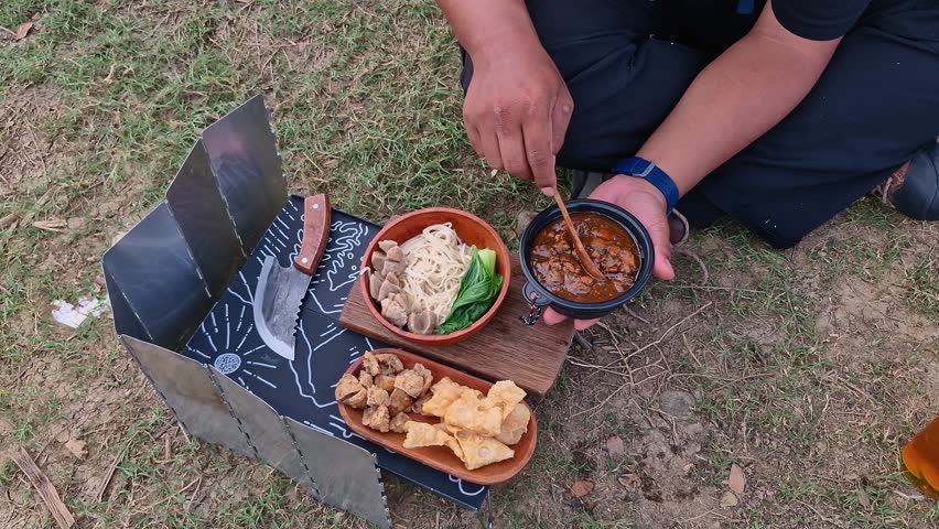 A male adventurer is preparing and cooking meatball chicken noodles on the edge of the reservoir. The concept of bushcraft camping on the edge of a lake and cooking with simple equipment