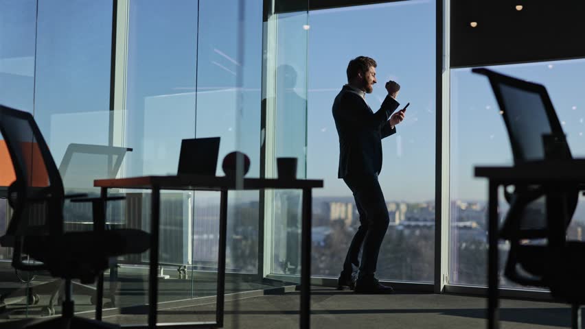 Excited businessman celebrates a triumph in a modern office. He holds phone, raises fist victoriously, showcasing success, motivation, and achievement.