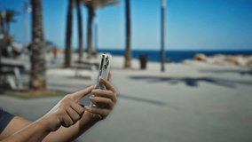 Man holding smartphone outdoors on sunny city street with palm trees and ocean in background, showcasing modern lifestyle and technology use. - Powered by Shutterstock - Get 15% off with code: PIKWIZARD15