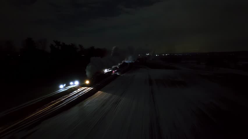 A steam locomotive moves steadily through a snow-covered area under the night sky, puffing clouds of steam. The soft glow of its headlights illuminates the surrounding landscape.