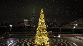 Yellow warm Christmas tree with snow flurries on pier with Manhattan, New York City skyline in background at night. Static aerial drone shot. - Powered by Shutterstock - Get 15% off with code: PIKWIZARD15