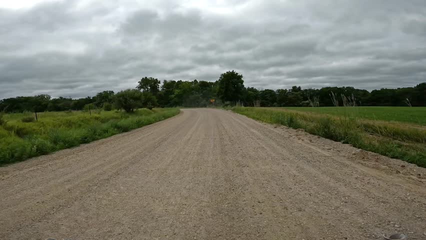 POV - driving on a curved gravel road; driving past corn fields and woods areas rural Iowa on a cloudy day; concepts of country life