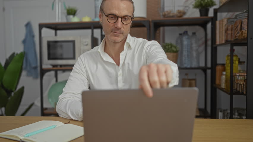 Mature caucasian man with glasses working on laptop in modern office setting with shelves and plants visible in the background - Powered by Shutterstock - Get 15% off with code: PIKWIZARD15