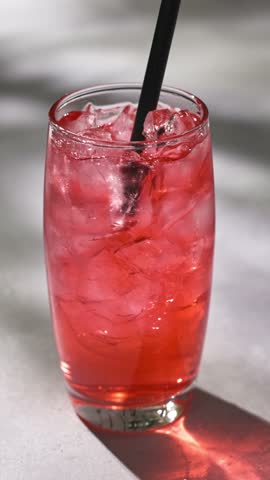 cocktail glass of fresh red drink with ice cubes on a gray sunny restaurant tabletop. hand put straw into glass