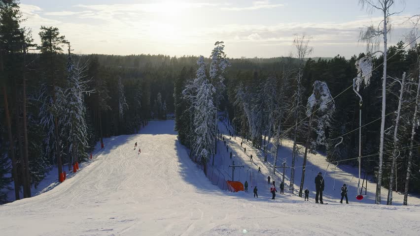 Skiers and snowboarders glide down a snowy slope surrounded by forest. A ski lift operates on the right, with sunlight casting long shadows.