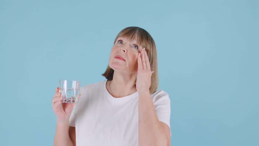 An elderly woman feels discomfort from pain in her head, swallows a pill and drinks it with water on a blue background in a calm indoor environment