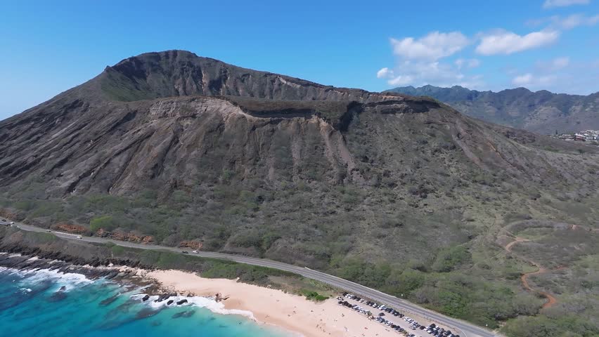 Aerial pan of Oahu, Hawaii, showing a coastal area with a mountain ridge, winding road, sandy beach, turquoise waters, and lush greenery.
