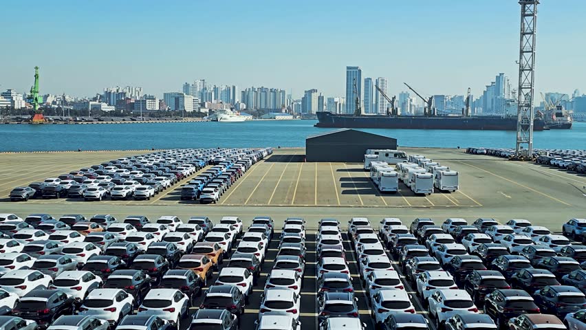 Thousands of brand-new cars for export are neatly parked at Wolmi Port, Incheon, with the city skyline and docked ships in the background under a clear blue sky - aerial dolly