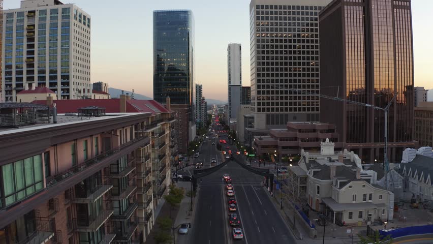 Aerial drone shot of downtown Salt Lake City, dollying out over State Street. Skyscrapers, apartments, and offices line the street, with fall trees glowing under a golden-orange autumn sunset.