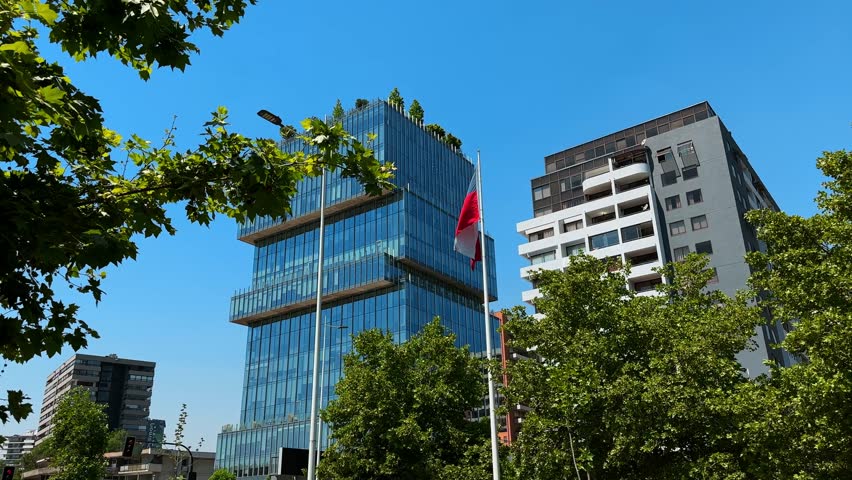 Static establishing shot of modern buildings in Santiago, Chile, on a clear day. The Chilean flag waves prominently in the wind, showcasing national pride against the urban skyline.