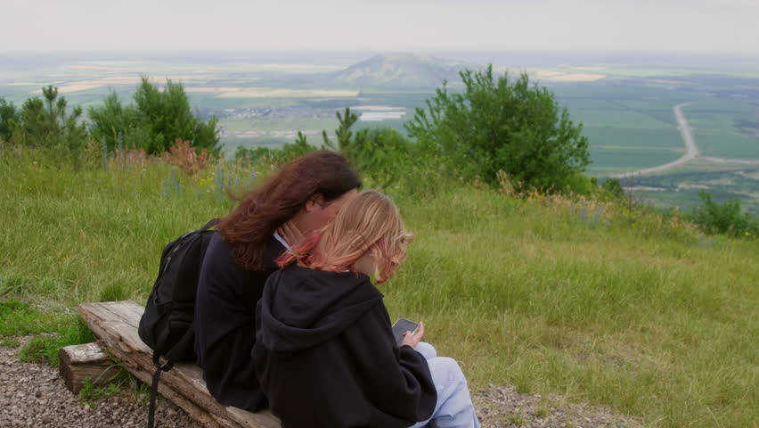 High angle view of mother and daughter looking at smartphone. Females wearing black jackets are sitting on bench over hill. They are exploring nature during windy day.