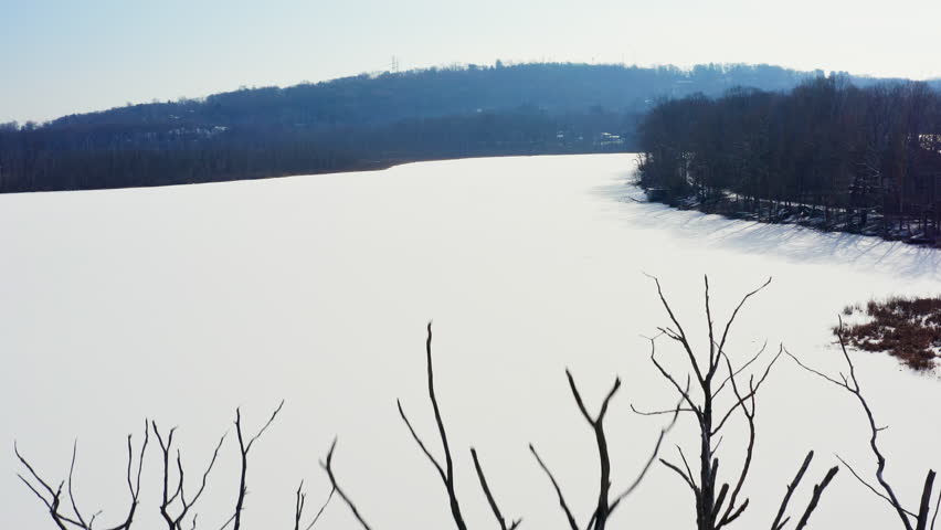 Aerial drone shot flying up over the frozen Mohansic Lake in FDR Park, Yorktown Heights, NY. Snow blankets the landscape as tree shadows stretch across the icy expanse.