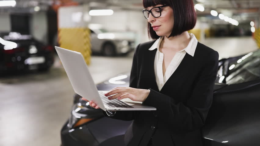 Caucasian businesswoman using laptop in indoor parking lot, focused on work. Woman wearing formal attire, glasses, standing near car. Professional and tech-savvy environment.