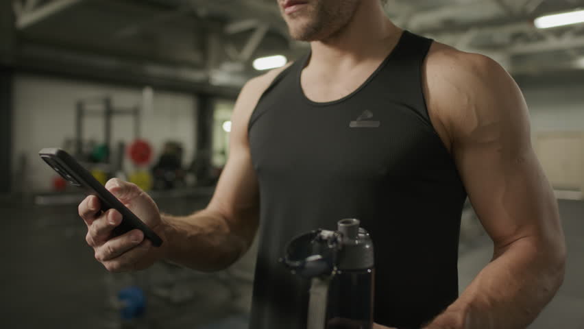 Muscular man in black tank top holding water bottle and scrolling on smartphone, taking break after workout in gym. Close-up view