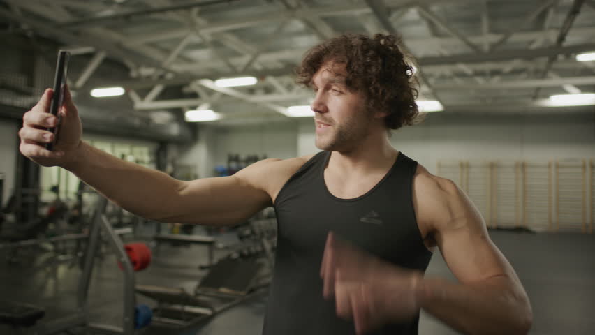 Muscular fitness blogger in sportswear standing in gym, waving and talking to smartphone camera during live stream on social media