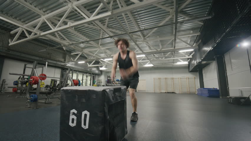 Muscular man in sportswear performing powerful box jumps in spacious gym during functional training session with padded plyometric box
