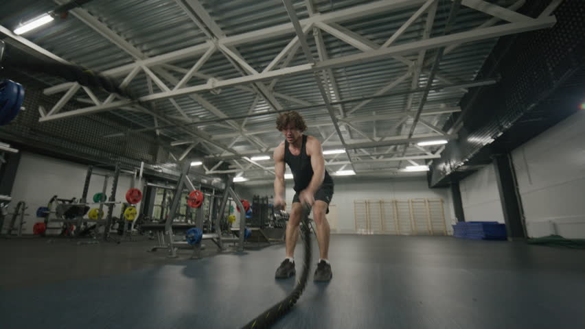 Strong muscular man doing intense battle rope exercise during training session in modern gym with weightlifting equipment. Zoom-in shot