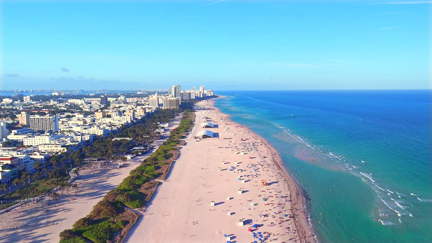 Drone hovers over the sand, rotating 360° to reveal South Beach in every direction—ocean waves, golden shoreline, Miami skyline, and the endless coastal view stretching north and south.