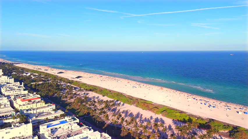 Drone captures a stunning 360° view over South Beach, Miami, revealing ocean waves, golden sand, South Pointe, the downtown skyline at sunset, and the coastline stretching north.