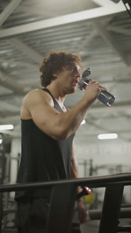 Young strong man in sportswear drinking water from bottle, then picking up pace and running on treadmill in gym. Vertical format footage