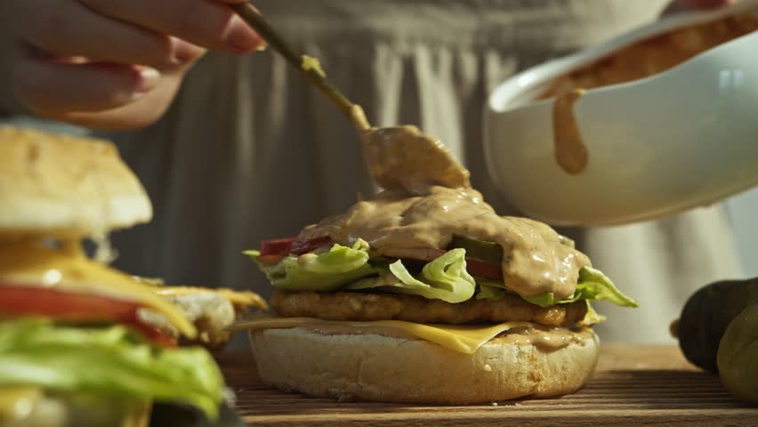Cook pours generous amount of sauce onto burger, based on mayonnaise and ketchup. This gives burger delicious, juicy taste, but it also has high calorie content, making it fatty junk food. Close up.