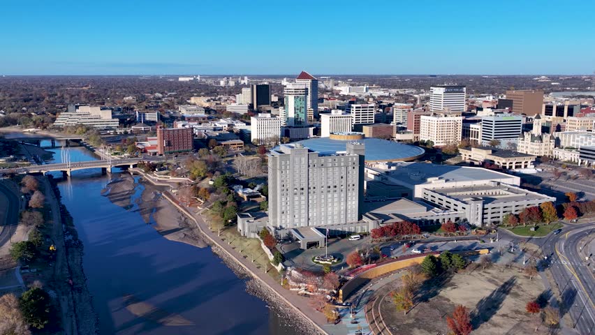 A rotating drone shot, flying high over Waterman Street, capturing a wide-angle view of the Arkansas River cutting through downtown Wichita, Kansas, revealing the uniquely shaped Arts Center building.