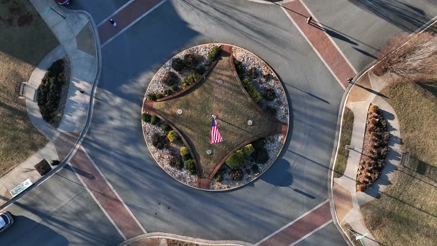 Flag of american waving on flagpole in center of roundabout. Pedestrians Crossing road at sunset time. Aerial top down circling shot. Cars entering and leaving roundabout in housing area.