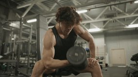 Strong man wearing black tank top performing one-arm bicep curls with dumbbell while sitting on bench in modern gym, surrounded by weightlifting equipment and machines - Powered by Shutterstock - Get 15% off with code: PIKWIZARD15