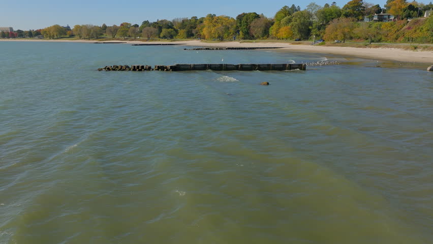 Aerial shot flying over blue waters of Lake Michigan, piers and a sandy beach in Sheboygan, Wisconsin in Fall. Gentle waves, coastal scenery, and a peaceful shoreline create a stunning lakeside view