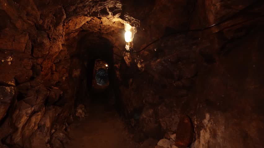 A dimly lit underground tunnel showcasing rugged rock walls, illuminated by faint lights, revealing the secrets of an ancient mine. (POV)