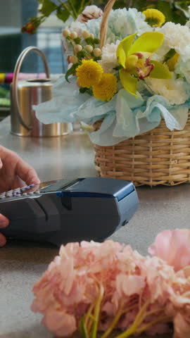 Vertical close-up view of hand of unknown female customer tapping card on payment terminal held by seller when buying flowers in shop