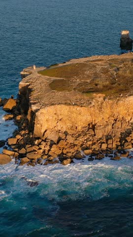 Aerial establishing shot of small rocky peninsula and ocean waves at sunset. Blue sea waves beat and splash in summer sunset haze, high cliffs and big stones, high edge of headland, Peniche, Portugal