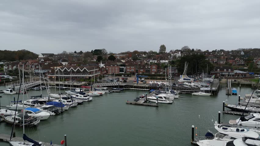 Drone shot of boats moored at harbor under cloudy sky on south coast of England, with water gently reflecting the boats and surrounding area.