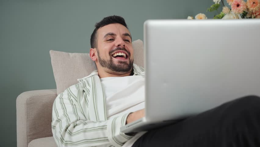 Joyful young man laughing heartily while using a laptop on a cozy sofa, enjoying a delightful moment filled with happiness and amusement in a relaxed indoor space, creating a cheerful leisure. High
