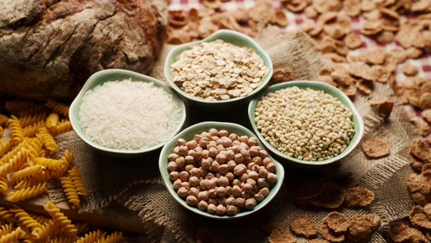 Close-up of carbohydrate-rich foods on a table, including bread, rice, wholegrain flakes, oats, wheat and corn pasta. Carbs Food.